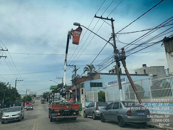 Homem está dentro de um cesto, erguido em um guindaste, fazendo troca de luminária em poste, numa rua. 
